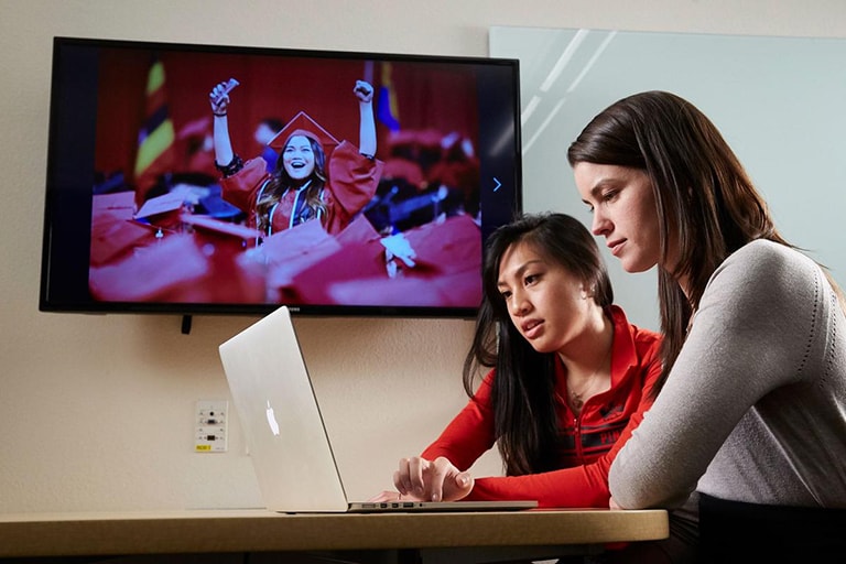 Two women working together on a laptop with an image of a UNLV grad on the TV behind them.